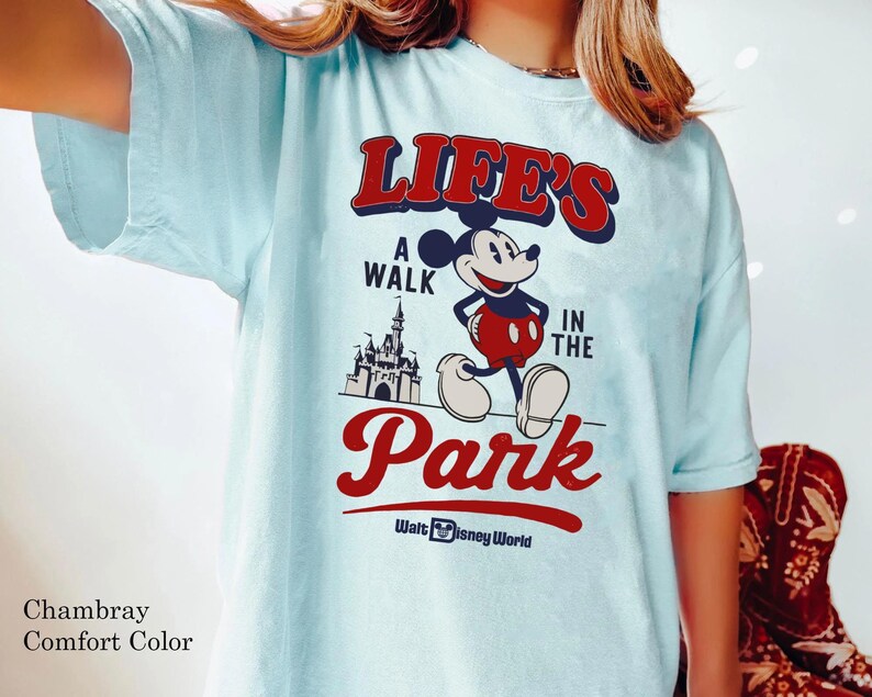 Puede incluir: Camiseta azul claro con un gr&aacute;fico de Mickey Mouse y el texto "LIFE'S A WALK IN THE PARK" en rojo y blanco. El dise&ntilde;o incluye un castillo y "Walt Disney World". La camiseta es de la marca Comfort Color.