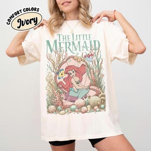 May include: Ivory-colored t-shirt featuring the text "The Little Mermaid" in green. The graphic depicts Ariel in a seashell with sea creatures and coral. The shirt has a relaxed fit and is made of a soft material.
