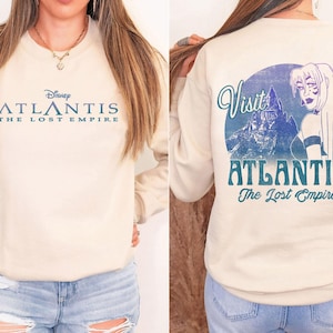 Könnte beinhalten: Cremefarbenes Sweatshirt mit dem Disney-Film "Atlantis: The Lost Empire". Auf der Vorderseite steht der Filmtitel in Blau. Auf der Rückseite befindet sich ein kreisförmiges Design mit den Worten "Visit Atlantis" und einer Charakterillustration.