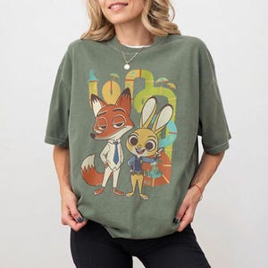 May include: Olive green t-shirt featuring a cartoon illustration of Nick Wilde and Judy Hopps from Zootopia. The graphic includes the characters in their signature outfits, set against a colorful, abstract background.