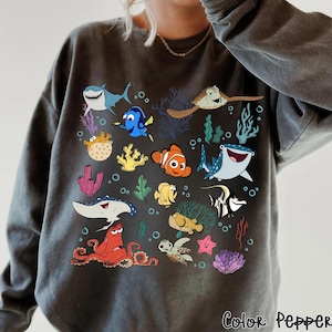 Può includere: Felpa grigio scuro con una grafica colorata di vari creature marine dei cartoni animati, tra cui squali, pesci e una tartaruga. Il design include coralli e bolle. Il testo "Color: Pepper" è stampato in basso a destra.