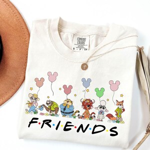 May include: Cream-colored t-shirt featuring a cartoon graphic of animal characters holding Mickey Mouse-shaped balloons. The word "FRIENDS" is printed in black. A gold necklace and a brown hat are also visible.