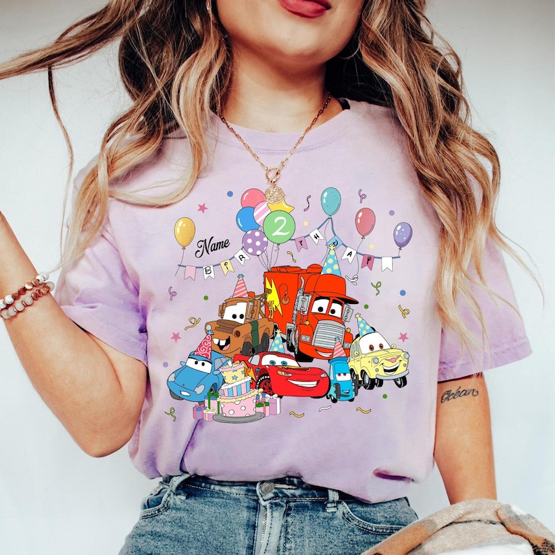May include: A light purple t-shirt featuring a colorful graphic of animated cars, balloons, and a birthday cake. The design includes the text "Name" and "2". The shirt is a short-sleeved crew neck.