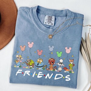 May include: A light blue Comfort Colors t-shirt featuring a cartoon graphic of animal characters holding Mickey Mouse-shaped balloons. The word "FRIENDS" is printed in white at the bottom. A gold necklace is draped across the shirt.