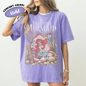 May include: Violet Comfort Colors t-shirt featuring a graphic of Ariel from The Little Mermaid. The design includes Ariel, Flounder, and Sebastian, surrounded by coral and seashells. The text "THE LITTLE MERMAID" is printed above the graphic. The shirt has a relaxed fit.