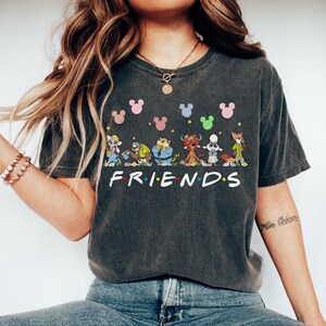 May include: Dark gray t-shirt featuring cartoon characters and Mickey Mouse balloons above the word "FRIENDS" in white. The shirt has a vintage wash appearance and a crew neck.