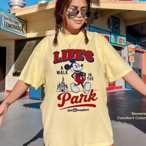 Puede incluir: Camiseta amarilla clara con el texto "LIFE'S A WALK IN THE PARK" y un dibujo de Mickey Mouse y un castillo. La camiseta tambi&eacute;n tiene el texto "Walt Disney World".