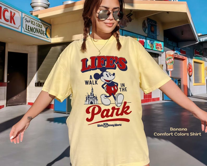 Puede incluir: Camiseta amarilla clara con el texto "LIFE'S A WALK IN THE PARK" y un dibujo de Mickey Mouse y un castillo. La camiseta tambi&eacute;n tiene el texto "Walt Disney World".