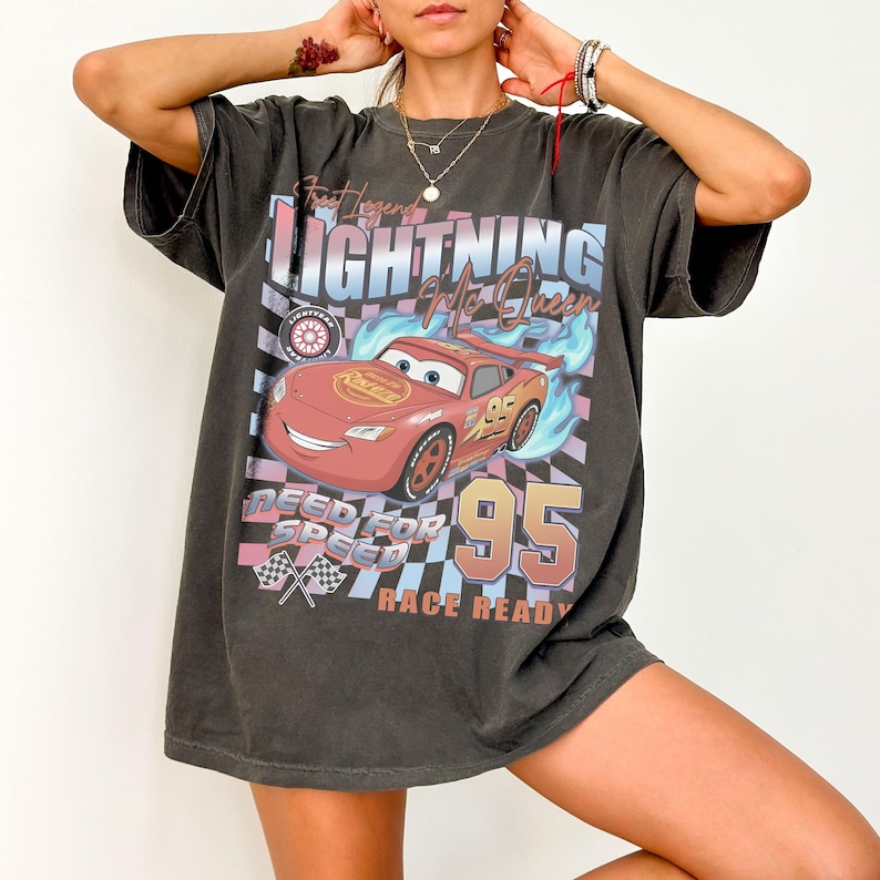 May include: Dark gray t-shirt featuring a graphic of Lightning McQueen from the movie Cars. The design includes the text "Lightning McQueen", "Need for Speed", and "Race Ready", along with the number "95" and checkered flags.