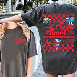 Puede incluir: Camiseta gris oscuro con el logo de Pizza Planet en rojo en la parte delantera y trasera. El dise&ntilde;o trasero incluye extraterrestres de dibujos animados, una hamburguesa y el texto "Friday Night Kids Night."