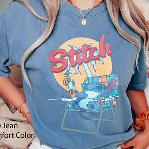 May include: Blue jean comfort color t-shirt featuring a cartoon Stitch character relaxing in a beach chair, wearing sunglasses, and holding a drink. The word "Stitch" is written in red and orange script above the character, with a palm tree and sun graphic.