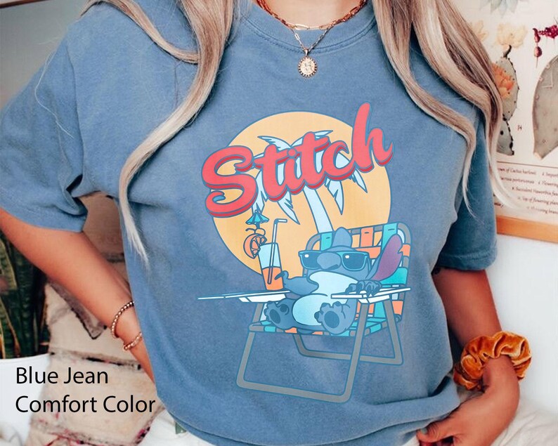 May include: Blue jean comfort color t-shirt featuring a cartoon Stitch character relaxing in a beach chair, wearing sunglasses, and holding a drink. The word "Stitch" is written in red and orange script above the character, with a palm tree and sun graphic.