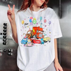 May include: White t-shirt featuring a colorful graphic of animated cars, balloons, and a birthday cake. The design includes the number "2" and the word "Name" above the cars, with a banner that says "BIRTHDAY".
