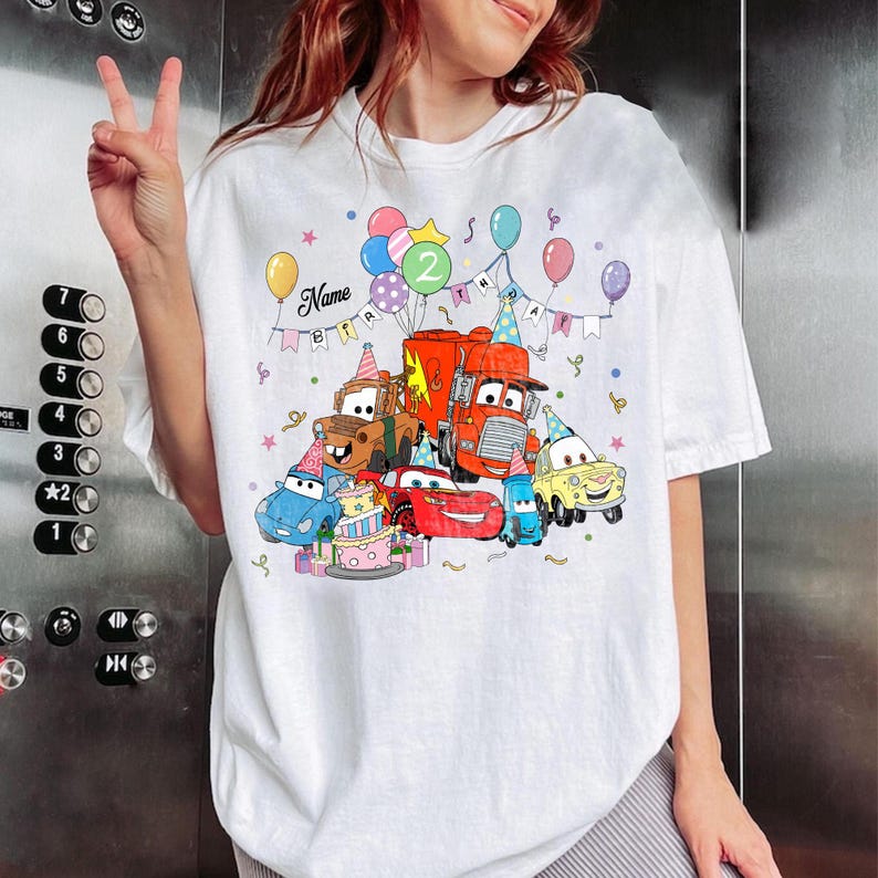 May include: White t-shirt featuring a colorful graphic of animated cars, balloons, and a birthday cake. The design includes the number "2" and the word "Name" above the cars, with a banner that says "BIRTHDAY".