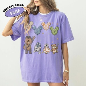 May include: A violet Comfort Colors t-shirt featuring Star Wars characters and Mickey Mouse-shaped balloons. The design includes Chewbacca, R2-D2, BB-8, and an Ewok. The balloons are in the shape of Mickey Mouse ears, with various patterns and colors.