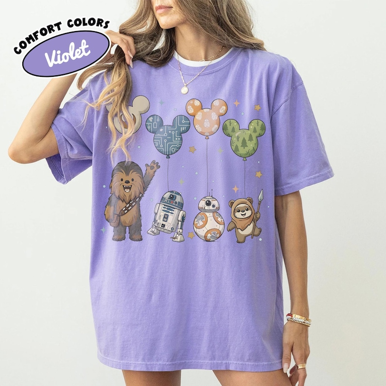 May include: A violet Comfort Colors t-shirt featuring Star Wars characters and Mickey Mouse-shaped balloons. The design includes Chewbacca, R2-D2, BB-8, and an Ewok. The balloons are in the shape of Mickey Mouse ears, with various patterns and colors.