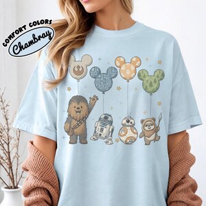 May include: Light blue t-shirt featuring cartoon characters from Star Wars, including Chewbacca, R2-D2, BB-8, and an Ewok. The characters are beneath Mickey Mouse-shaped balloons. The shirt has the text "COMFORT COLORS Chambray".