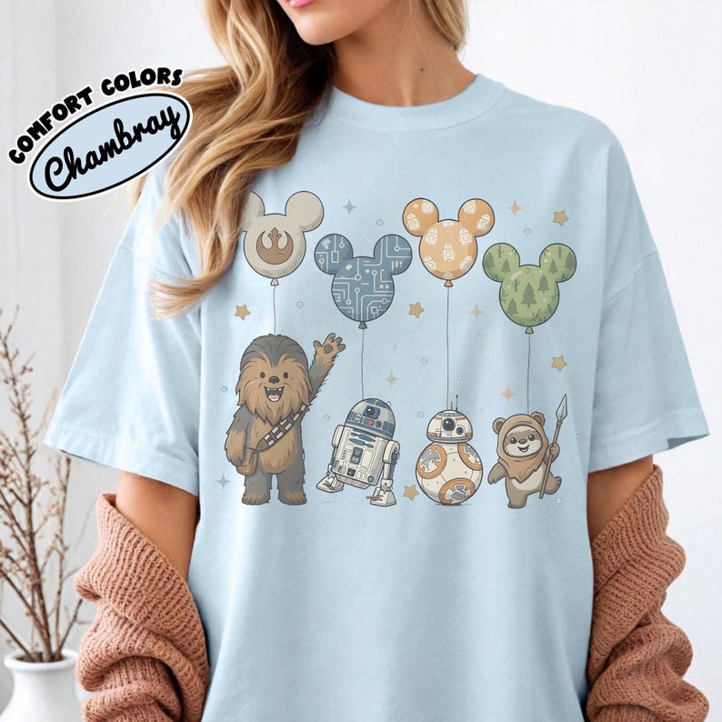 May include: Light blue t-shirt featuring cartoon characters from Star Wars, including Chewbacca, R2-D2, BB-8, and an Ewok. The characters are beneath Mickey Mouse-shaped balloons. The shirt has the text "COMFORT COLORS Chambray".