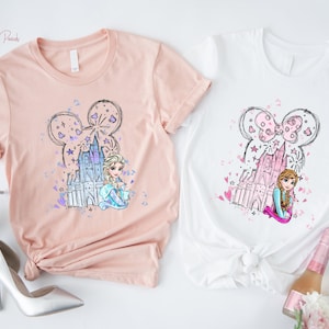 May include: Two t-shirts, one peach and one white, featuring a cartoon design. The design includes a castle, stars, and a character's face within a Mickey Mouse ear outline. The peach shirt has the text "Heather Peach". The white shirt has the text "White".