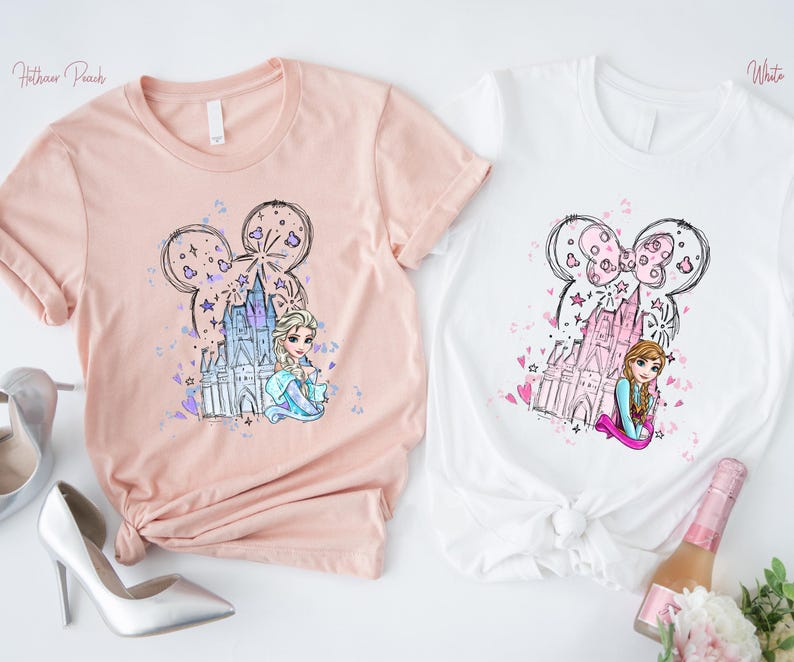May include: Two t-shirts, one peach and one white, featuring a cartoon design. The design includes a castle, stars, and a character's face within a Mickey Mouse ear outline. The peach shirt has the text "Heather Peach". The white shirt has the text "White".