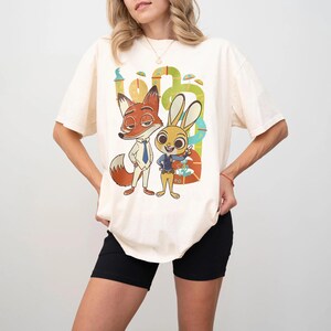May include: Cream-colored t-shirt featuring a colorful graphic of the Zootopia characters Nick Wilde and Judy Hopps. The cartoon fox and bunny are standing in front of a green and yellow background. The shirt has short sleeves.