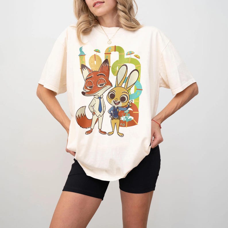 May include: Cream-colored t-shirt featuring a colorful graphic of the Zootopia characters Nick Wilde and Judy Hopps. The cartoon fox and bunny are standing in front of a green and yellow background. The shirt has short sleeves.