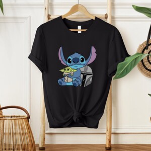 May include: Black t-shirt featuring a cartoon illustration of Stitch holding Grogu, with a silver helmet. The design includes blue, green, and gray colors. The shirt is hanging on a wooden hanger.