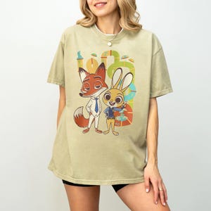 May include: A light green t-shirt featuring cartoon characters Nick Wilde the fox and Judy Hopps the rabbit from Zootopia. The graphic includes the characters in their outfits, with a colorful background. The shirt is a relaxed fit.