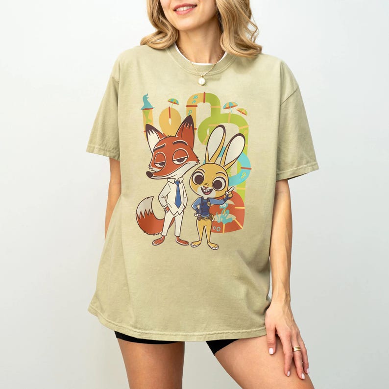 May include: A light green t-shirt featuring cartoon characters Nick Wilde the fox and Judy Hopps the rabbit from Zootopia. The graphic includes the characters in their outfits, with a colorful background. The shirt is a relaxed fit.