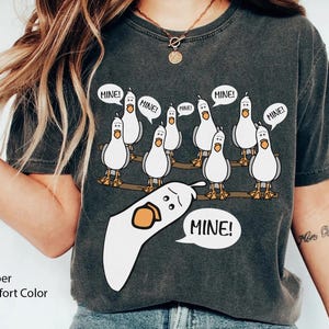 May include: Pepper Comfort Color t-shirt featuring a cartoon of seagulls on a wooden beam, each with a speech bubble saying "MINE!". One seagull is upside down with a speech bubble saying "MINE!".