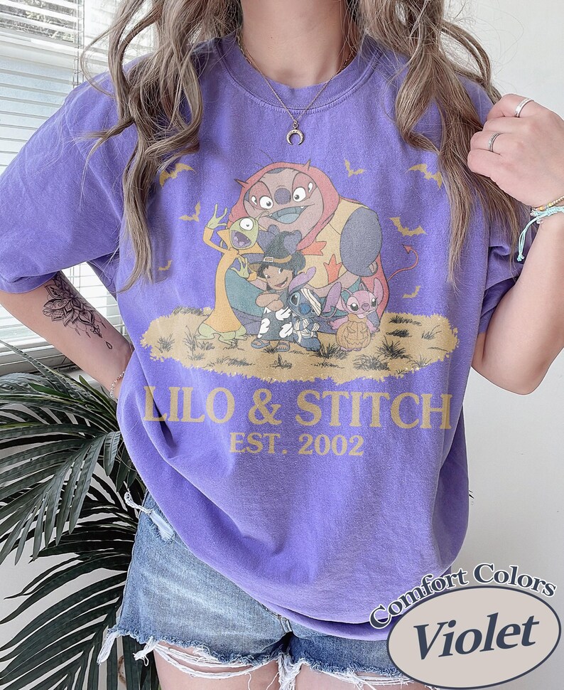 Op de afbeelding: Violet Comfort Colors T-shirt met een Lilo & Stitch-afbeelding. Het ontwerp bevat de tekst "Lilo & Stitch EST. 2002" in geel. Het shirt heeft een effen violette kleur.