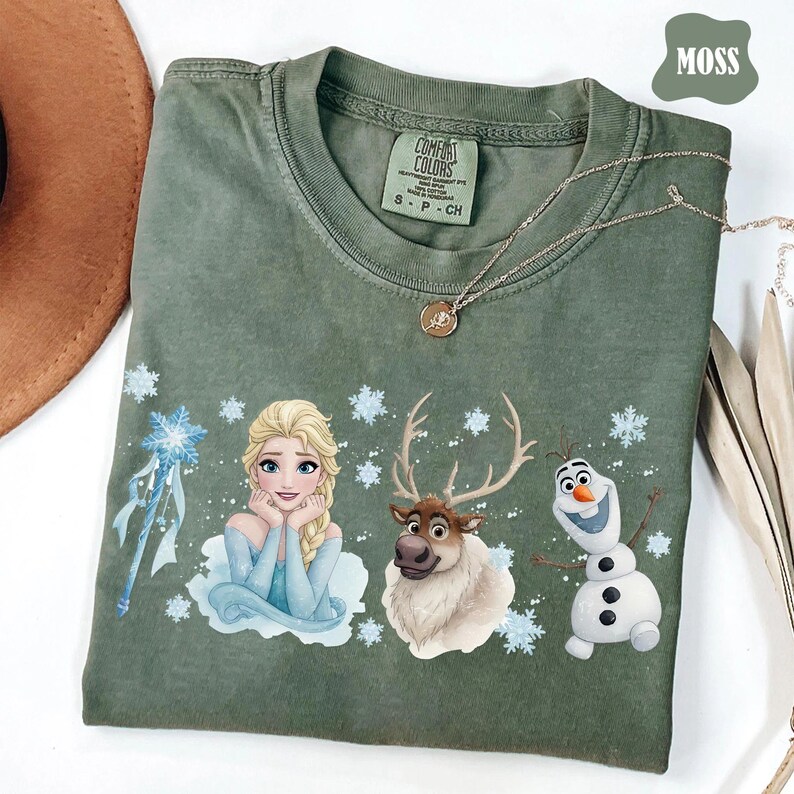 Peut inclure: T-shirt vert mousse avec un motif d'Elsa, Sven et Olaf du film La Reine des Neiges. Le t-shirt a un col rond et des manches courtes. Un collier argent&eacute; avec un pendentif est drap&eacute; sur le t-shirt.