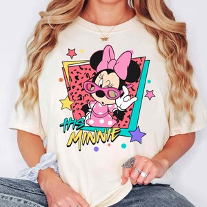 K&ouml;nnte beinhalten: Cremefarbenes T-Shirt mit einer Grafik von Minnie Mouse mit rosa Sonnenbrille und einem gepunkteten Kleid. Das Design enth&auml;lt den Text "His Minnie" in einem Retro-Stil mit bunten Sternen und geometrischen Formen.