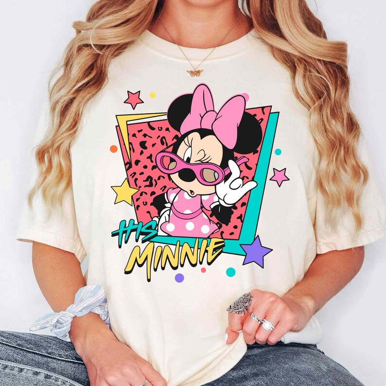 K&ouml;nnte beinhalten: Cremefarbenes T-Shirt mit einer Grafik von Minnie Mouse mit rosa Sonnenbrille und einem gepunkteten Kleid. Das Design enth&auml;lt den Text "His Minnie" in einem Retro-Stil mit bunten Sternen und geometrischen Formen.