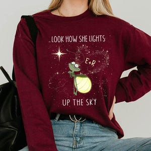May include: Burgundy long-sleeve sweatshirt with a graphic of a firefly holding a glowing lantern. The text reads, ".LOOK HOW SHE LIGHTS UP THE SKY." The firefly is next to a star and a heart-shaped constellation.