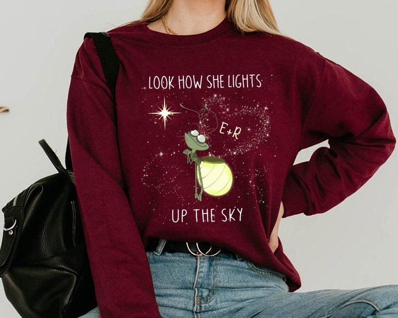 May include: Burgundy long-sleeve sweatshirt with a graphic of a firefly holding a glowing lantern. The text reads, ".LOOK HOW SHE LIGHTS UP THE SKY." The firefly is next to a star and a heart-shaped constellation.