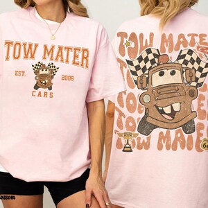 May include: Pink t-shirt featuring a graphic of Tow Mater from the Cars movie. The front of the shirt displays "Tow Mater" in orange lettering, with "Est. 2006" and "Cars" below. The back features a larger image of Mater with checkered flags.