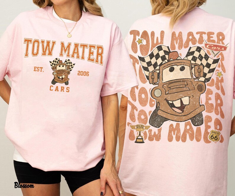 May include: Pink t-shirt featuring a graphic of Tow Mater from the Cars movie. The front of the shirt displays "Tow Mater" in orange lettering, with "Est. 2006" and "Cars" below. The back features a larger image of Mater with checkered flags.