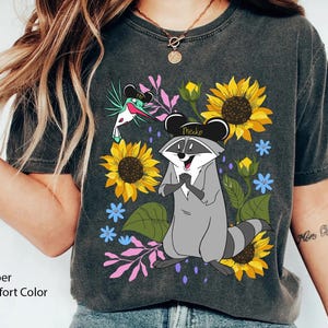 Puede incluir: Una camiseta gris oscuro con un mapache de dibujos animados que lleva un sombrero negro con el nombre "Meeko", rodeado de girasoles, flores azules y un colibrí. La camiseta es de la marca Comfort Color.