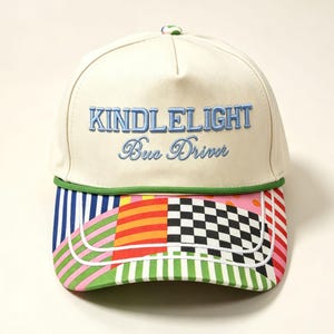 Puede incluir: Una gorra de béisbol color crema con el texto bordado KINDLELIGHT Bus Driver en azul claro. La visera presenta un patrón de patchwork multicolor que incluye rayas, lunares y un diseño de tablero de ajedrez en blanco y negro.