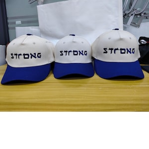 May include: Three baseball caps with off-white crowns and blue brims. Each cap features the word STRONG embroidered in dark blue, stylized lettering across the front panel. The caps have a structured design with a button on the top center.