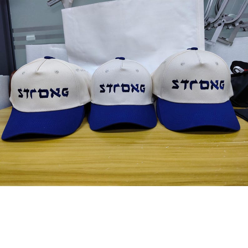 May include: Three baseball caps with off-white crowns and blue brims. Each cap features the word STRONG embroidered in dark blue, stylized lettering across the front panel. The caps have a structured design with a button on the top center.