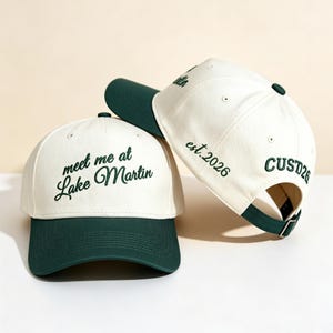 Puede incluir: Dos gorras de béisbol con corona color crema y visera verde oscuro. La gorra frontal muestra el texto bordado "meet me at Lake Martin" en verde oscuro. La gorra lateral presenta el texto bordado "est.2026" y "CUSTD26".