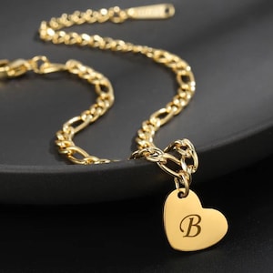 Puede incluir: Collar dorado con un colgante en forma de corazón. El colgante tiene grabada la letra "B". La cadena tiene un diseño de eslabones figaro. La joya se muestra sobre una superficie oscura.