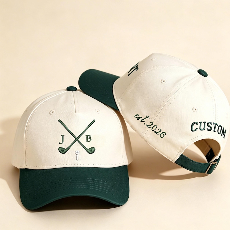 May include: Two cream and dark green baseball caps. The front cap features embroidered crossed golf clubs with the letters J and B and a golf tee. The side cap displays the embroidered text est. 2026 and the word CUSTOM in dark green thread.