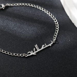 May include: A silver chain bracelet with a nameplate charm featuring the Arabic word "Jalil" in a simple, modern font.