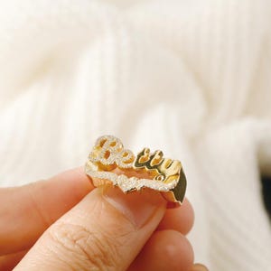 Könnte beinhalten: Goldring mit dem Namen "Becky" in Kursivschrift mit kleinen, klaren Steinen, die in die Buchstaben eingefasst sind.