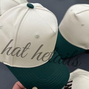 May include: A five-panel baseball cap featuring a cream-colored crown and a dark green brim. The cap includes a matching dark green button at the top center of the crown. The design is structured with visible stitching along the panels and brim.