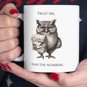 May include: White ceramic mug featuring a detailed illustration of an owl wearing glasses and holding a stack of papers. The mug has the text "TRUST ME, I RAN THE NUMBERS."