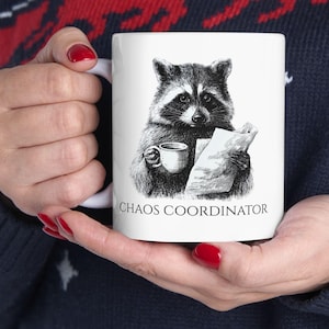 May include: White ceramic mug featuring a detailed black and white illustration of a raccoon holding a cup and papers. The text "CHAOS COORDINATOR" is printed below the raccoon. The mug is held by a person wearing a dark blue sweater.